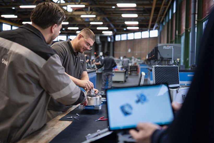Ausbildung bei thyssenkrupp Steel: links stehen zwei Azubis in Kluft an der Werkbank, im Vordergrund ist unscharf ein Tablet-Display zu sehen