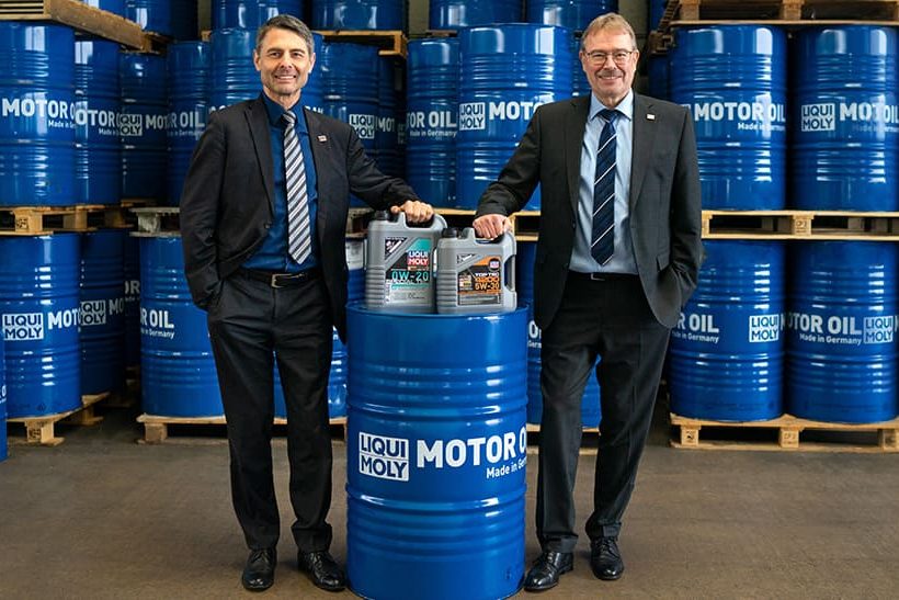 Zu sehen sind zwei weiße Männer im Anzug (die Geschäftsführer von Liqui Moly), die mit einer blauen Tonne und vor einer Wand aus blauen Tonnen posieren.