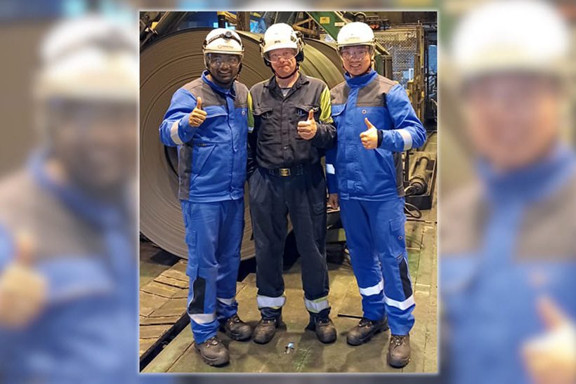 Das Bild zeigt drei Männer im Blaumann: Premsingh Bist, Projektleiter und leitender Ingenieur bei Primetals Technologies, Jari Puurtinen, Entwicklungsleiter für Prozessautomatisierung und Projektleiter für Elektrik und Automatisierung bei SSAB Hämeenlinna, und Dong Yan, Ingenieur für die Inbetriebnahme von Antrieben bei Primetals Technologies, vor Ort, als das erste Coil, nach Abschluss von Phase 1 der Modernisierung des Walzgerüsts bei SSAB Hämeenlinna, produziert wurde.