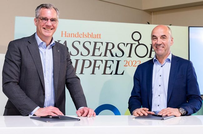 Das Bild zeigt zwei Männer mittleren Alters ab der Hüfte aufwärts, die Sakkos tragen und an einem Tisch stehen. Es sind Gunnar Groebler (Salzgitter AG, links) und Stefan Dohler (EWE). Die beiden lächeln in Richtung des Betrachters, da sie gerade eine Kooperationsvereinbarung für Wasserstoff abgeschlossen haben.