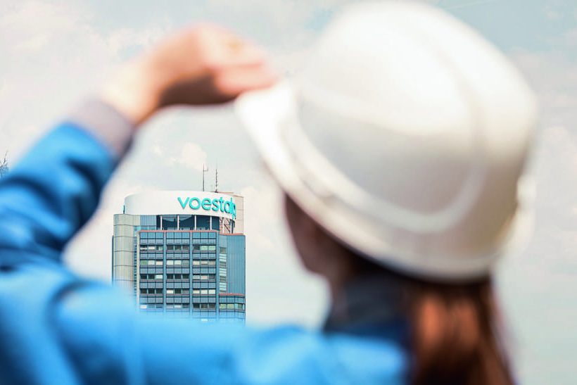 Das Bild zeigt im Vordergrund (und unscharf) eine weibliche Person von hinten, die ihre linke Hand an einen weißen Sicherheitshelm legt. Durch ihre Arme hindurch ist ein Bürogebäude von Voestalpine zu sehen. Zum Halbjahr 2024/25 liegt das Unternehmen unter der Markterwartung, erwartet aber schwarze Zahlen.