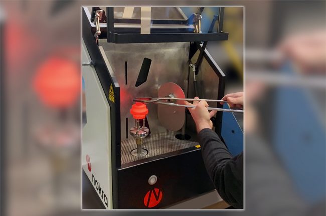 Nokra (web) Das Bild zeigt, wie ein warmes Schmiedestück unmittelbar nach dem Verlassen der Schmiedepresse direkt vom Förderband in eine Messzelle von Nokra eingesetzt wird.