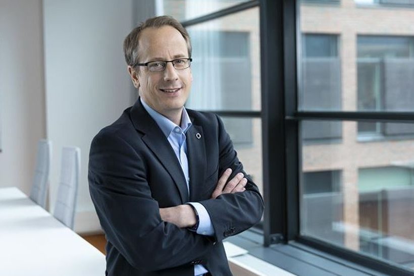 Das Bild zeigt einen Mann, der ein Sakko trägt und auf einem leeren Tisch sitzt. Es handelt sich um Marc-Simon Schaar von Outokumpu.