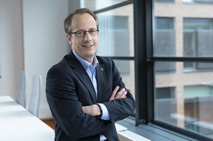 Das Bild zeigt einen Mann, der ein Sakko trägt und auf einem leeren Tisch sitzt. Es handelt sich um Marc-Simon Schaar von Outokumpu.