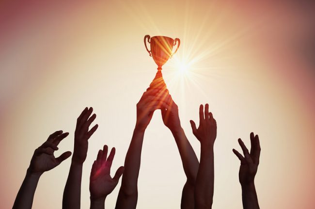 Das bild zeigt im Gegenlicht sieben in die Höhe gereckte Unterarme. Ein paar Hände hält dabei einen Pokal fest. Das Bild steht sinnbildlich für die jüngste Auszeichnung von Dillinger.