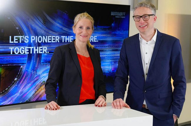 Das Bild zeigt eine weiße Frau (Birgit Held) und einen weißen Mann (Stephan Knüttel), die an einem Stehtisch stehen. Sie tragen Business-Kleidung und gehören zum Unternehmen Benteler.