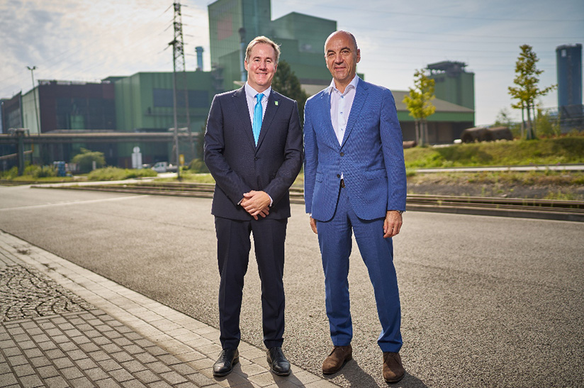 Das Bild zeigt zwei anzugtragende Männer im Vordergrund und eine Industrieanlage im Hintergrund. Es sind Dr. Alexander Becker (link,s dunkler Anzug), CEO der GMH Gruppe, und Stefan Dohler (rechts, blauer Anzug), EWE-Vorstands­vorsitzender.