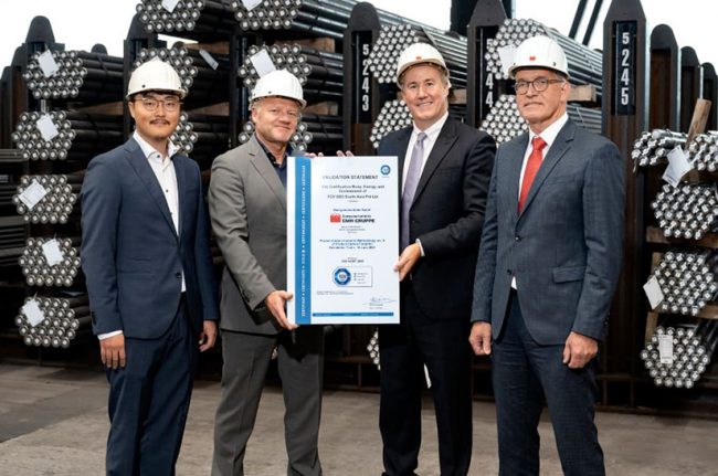 GMH Gruppe_TUV Certificate – conference_2023 (web) Das Bild zeigt vier männliche Anzugträger mit weißem Helmen, die in einem Lager mit Stahl stehen. Sie lächeln in die Kamera und die beiden in der Mitte halten gemeinsam eine große Urkunde. Es handelt sich hier um das Führungspersonal der GMH Gruppe und von TÜV Süd.