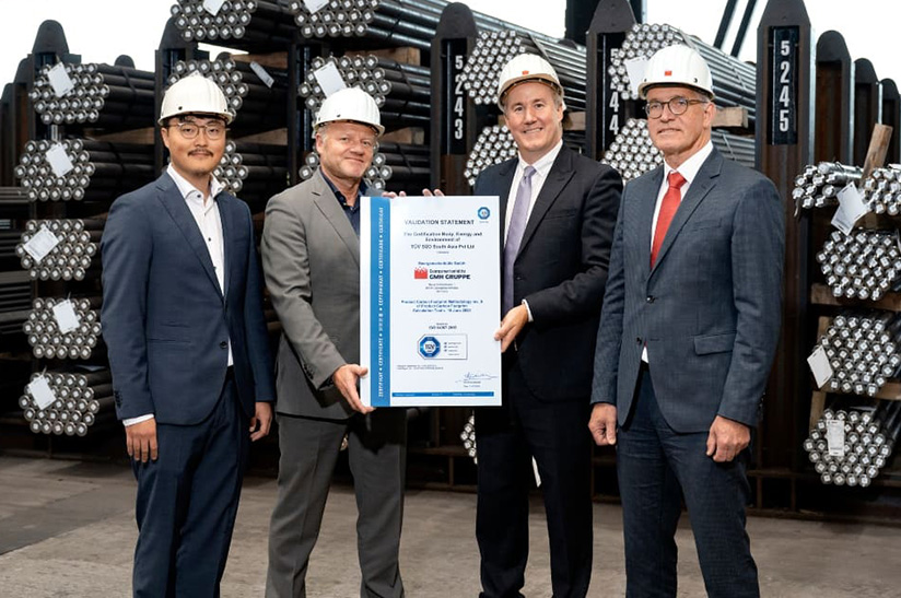 GMH Gruppe_TUV Certificate – conference_2023 (web) Das Bild zeigt vier männliche Anzugträger mit weißem Helmen, die in einem Lager mit Stahl stehen. Sie lächeln in die Kamera und die beiden in der Mitte halten gemeinsam eine große Urkunde. Es handelt sich hier um das Führungspersonal der GMH Gruppe und von TÜV Süd.