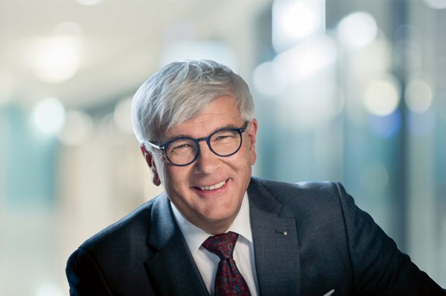 Noebauer (web) Das Bild zeigt das Porträt eines weißen, grauhaarigen Mannes. Er trägt eine Brille, einen Anzug mit Krawatte und lächelt in die Kamera. Es handelt sich dabei um Reinhard Noebauer von Voestalpine.