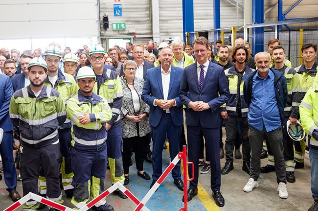 Das Bild zeigt eine Gruppe von Menschen bei der Einweihung einer neuen Anlage bei Outokumpu. In der ersten Reihe stelehen viele Mitarbeiter in Arbeitskluft und mit Helmen sowie NRW-Ministerpräsident Hendrik Wüst.