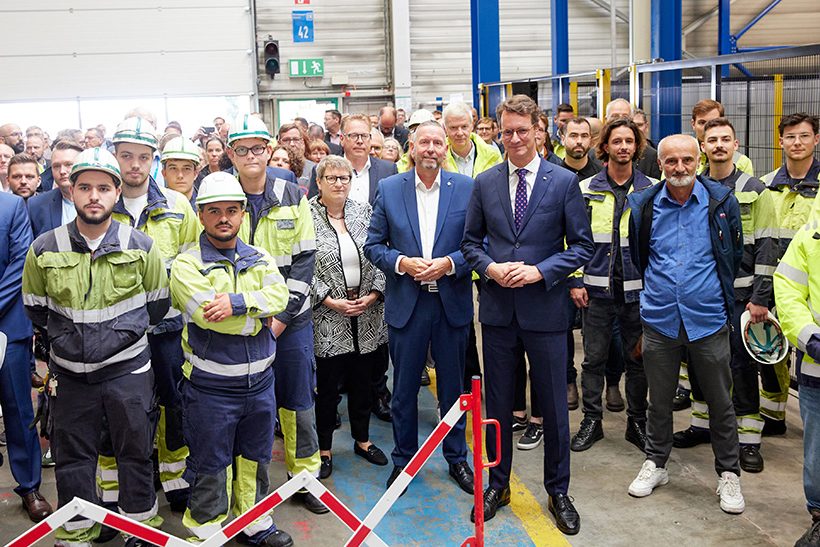 Das Bild zeigt eine Gruppe von Menschen bei der Einweihung einer neuen Anlage bei Outokumpu. In der ersten Reihe stelehen viele Mitarbeiter in Arbeitskluft und mit Helmen sowie NRW-Ministerpräsident Hendrik Wüst.