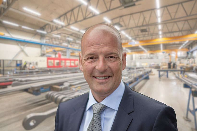 Das Bild zeigt einen Mann im Anzug (den Geschäftsführer von Proplate). Im Hintergrund ist unscharf eine Lagerhalle von innen zu sehen.