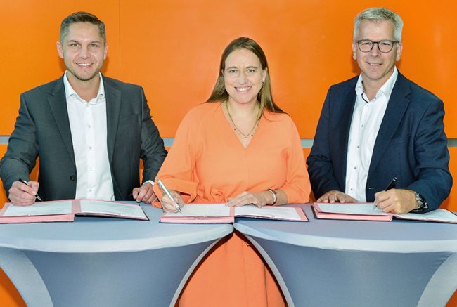 Das Bild zeigt drei Menschen, die an Stehtischen einen Vertrag unterzeichnen, um eine nachhaltige Logistik zu fördern. In der Mitte steht eine Frau in einem orangefarbenem Kleid, ihr zur Seite zwei Männer in Sakkos und weißen Hemden. Der Hintergrund ist ebenfalls orange.
