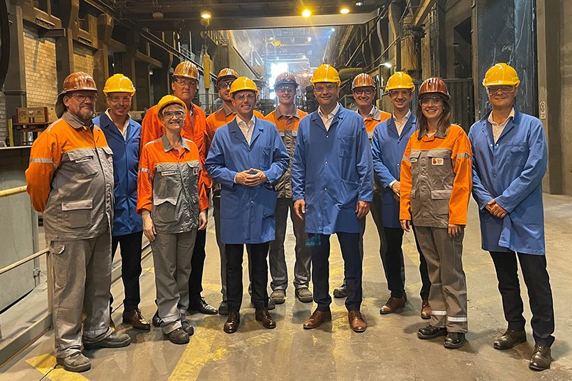 Das Bild zeigt eine Gruppe von Menschen im stahlwerk-szene-transformationsgipfel-aufruf der Swiss Steel Group. Mittig mit blauen Kitteln stehen links der US-Botschafter Scott Miller und der Luzerner Regierungspräsident Fabian Peter. Die Personen in orange-grauer Sicherheitskleidung gehören zum Unternehmen.