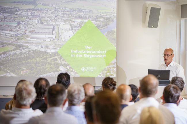 230911-ESF-Parlamentarischer-Abend-12_©Matthias_Seifert (web) Bei Feralpi Stahl fand ein Parlamentarischer Abend statt. Das Bild zeigt eine Vortragssituation: die Köpfe der sitzenden Zuhörer in der Rückansicht, rechts am Rednerpult einen Redner und an der Wand eine Präsentation.