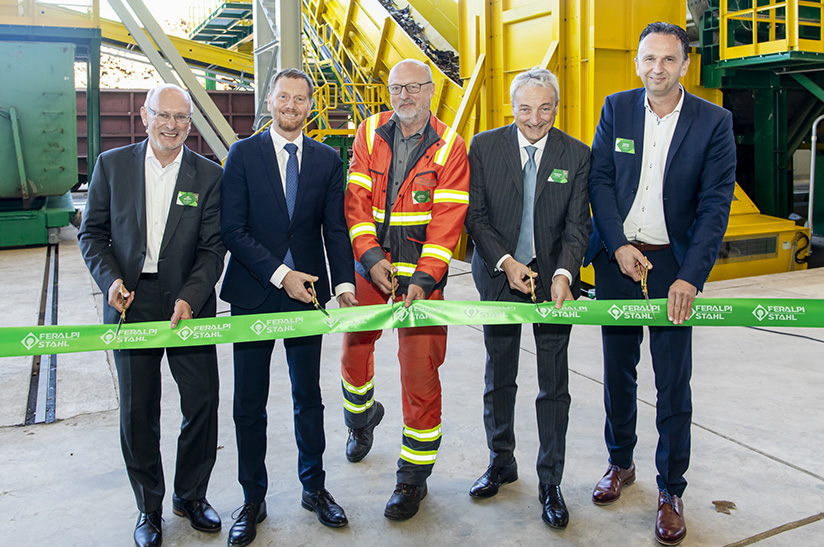 Schrottaufbereitung_Einweihung-Schrotthalle-ESF-258 (web) Das Bild zeigt vier Anzugträger und eine Person in Schutzausrüstung, die gemeinsam ein grünes Band zur Einweihung einer Schrottaufbereitung durchtrennen.