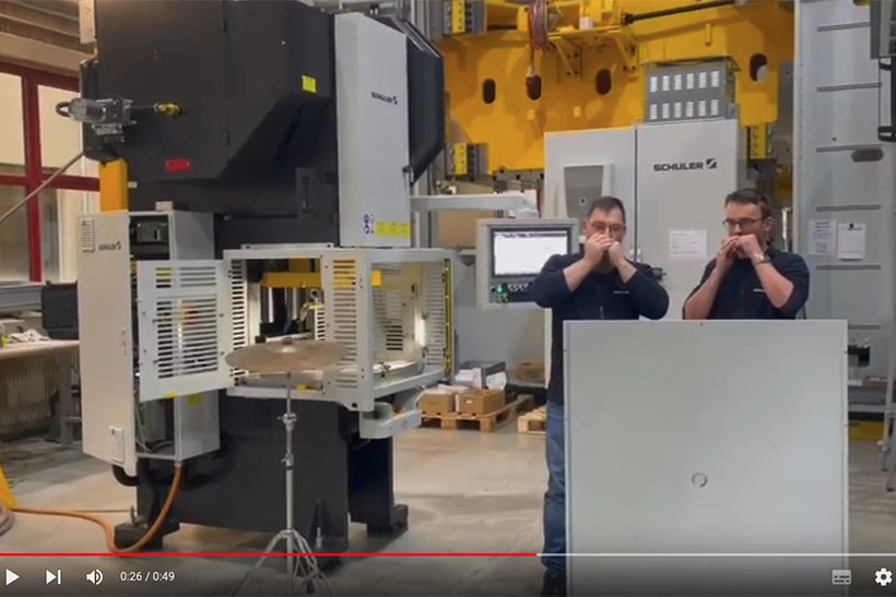 Das Bild zeigt einen Ausschnitt aus einem YouTube-Video der Schuler Group. Zwei Mitarbeiter spielen Mundharmonika und im Hintergrund hämmert eine Maschine auf ein Becken.