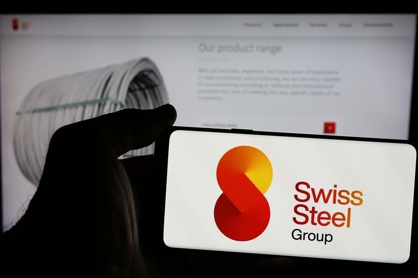 Das Bild zeigt das Logo der Swiss Steel Group (als Teil der Swiss Steel Holding) auf einem Smartphone-Display. Dahinte ist ein Monitor mit der Website des Unternehmens zu sehen.