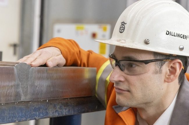 Stahlspezialist Dillinger hat im Forschungsprojekt HL-Blech (HochLeistungs-Blech) mit Verbundpartnern neue Stahlsorten entwickelt. Das Bild zeigt einen Mitarbeiter von Dillinger, der einen weißen Helm trägt und auf Stahl schaut. Dabei handelt es sich um Projektleiter Dr. Sebastian Scholl vor einer Hochenergie-Unterpulverschweißverbindung mit 7 kJ/mm zwecks Schweißeignungsprüfung an einem neuentwickelten Stahl der Güte S355ML mit einer Blechdicke von 80 mm.