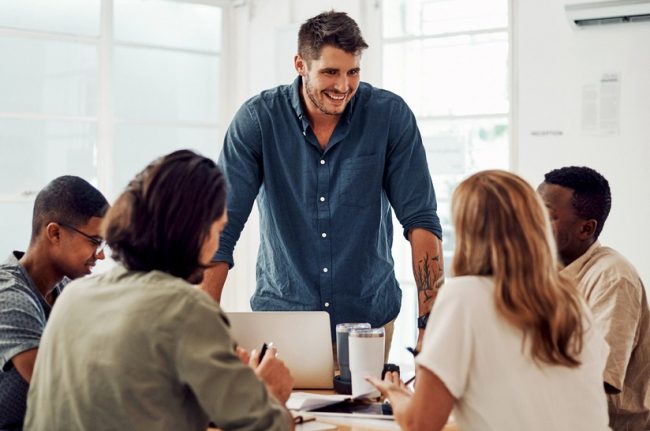 Teamwork,,Meeting,And,Manager,For,Training,In,Office,,Coworking,And Das Foto zeigt fünf junge Erwachsene in einer Besprechung. Vier sitzen und hören zu, einer steht und spricht. Es geht um gewaltfreie Kommunikation, entsprechend lächelt der Sprecher.