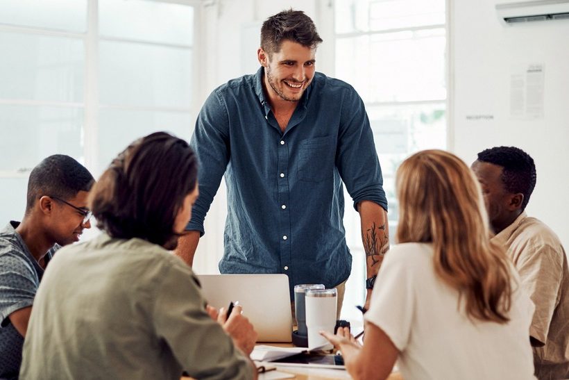 Das Foto zeigt fünf junge Erwachsene in einer Besprechung. Vier sitzen und hören zu, einer steht und spricht. Es geht um gewaltfreie Kommunikation, entsprechend lächelt der Sprecher.