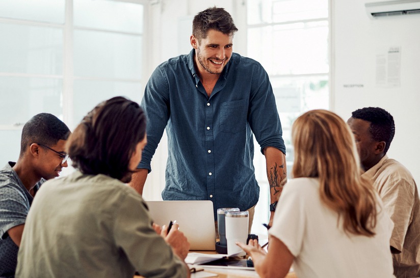 Teamwork,,Meeting,And,Manager,For,Training,In,Office,,Coworking,And Das Foto zeigt fünf junge Erwachsene in einer Besprechung. Vier sitzen und hören zu, einer steht und spricht. Es geht um gewaltfreie Kommunikation, entsprechend lächelt der Sprecher.